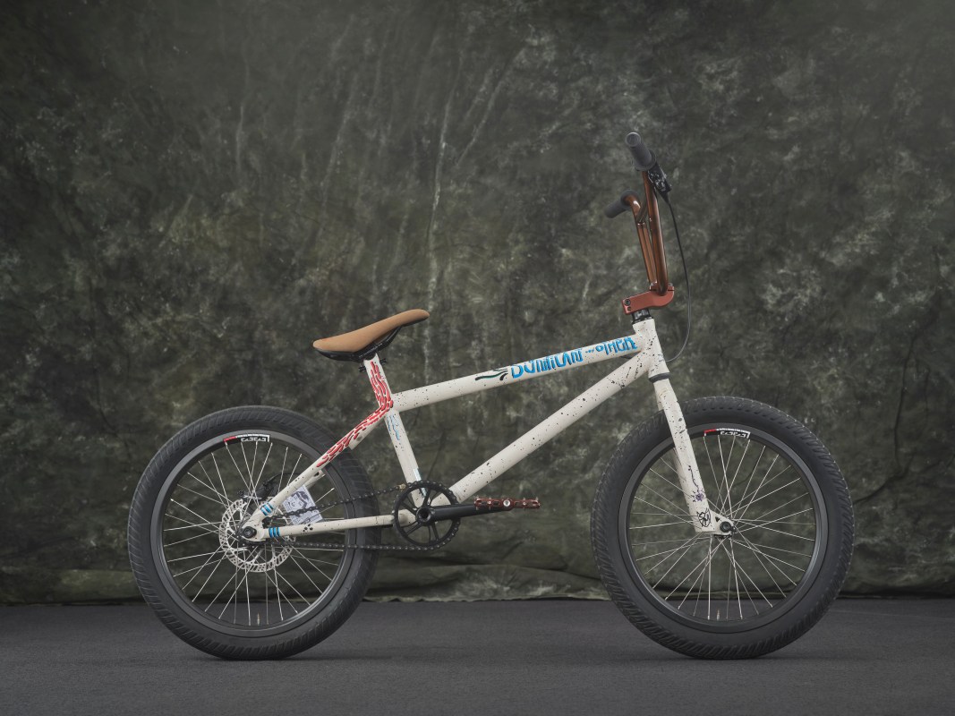 A custom bicycle featuring a white frame with colorful accents and a brown seat, positioned against a textured dark backdrop.