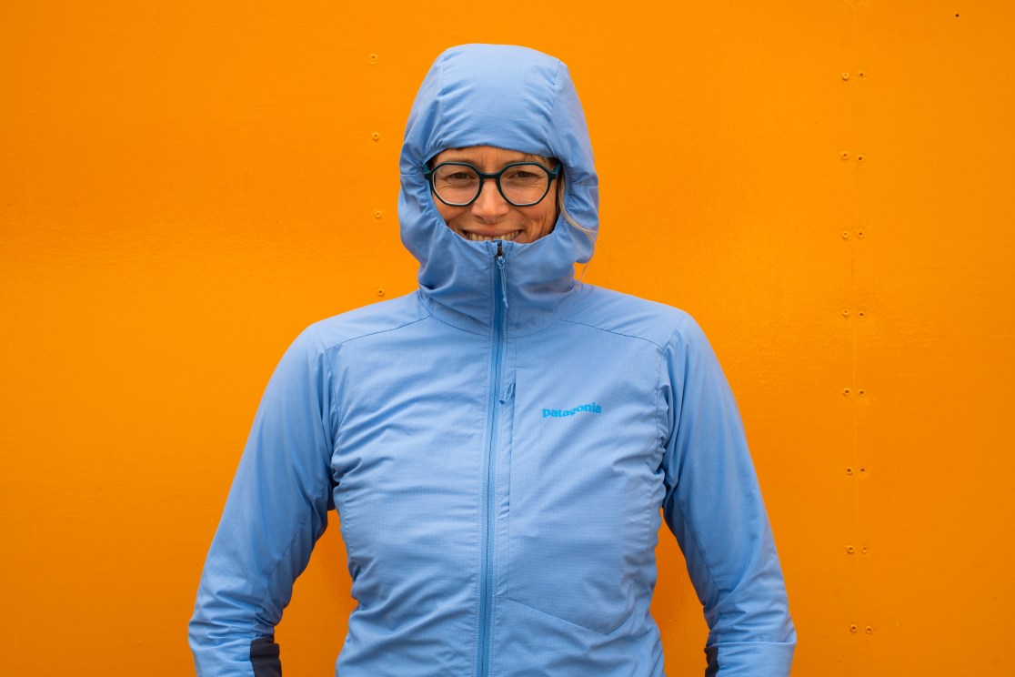 Person wearing a blue weather-resistant jacket with a hood, smiling in front of a bright orange background.
