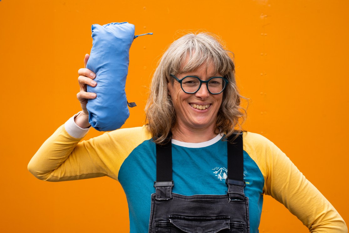 A person smiling while holding a blue packed item. They are wearing a colorful long-sleeve shirt and black overalls, with an orange background.