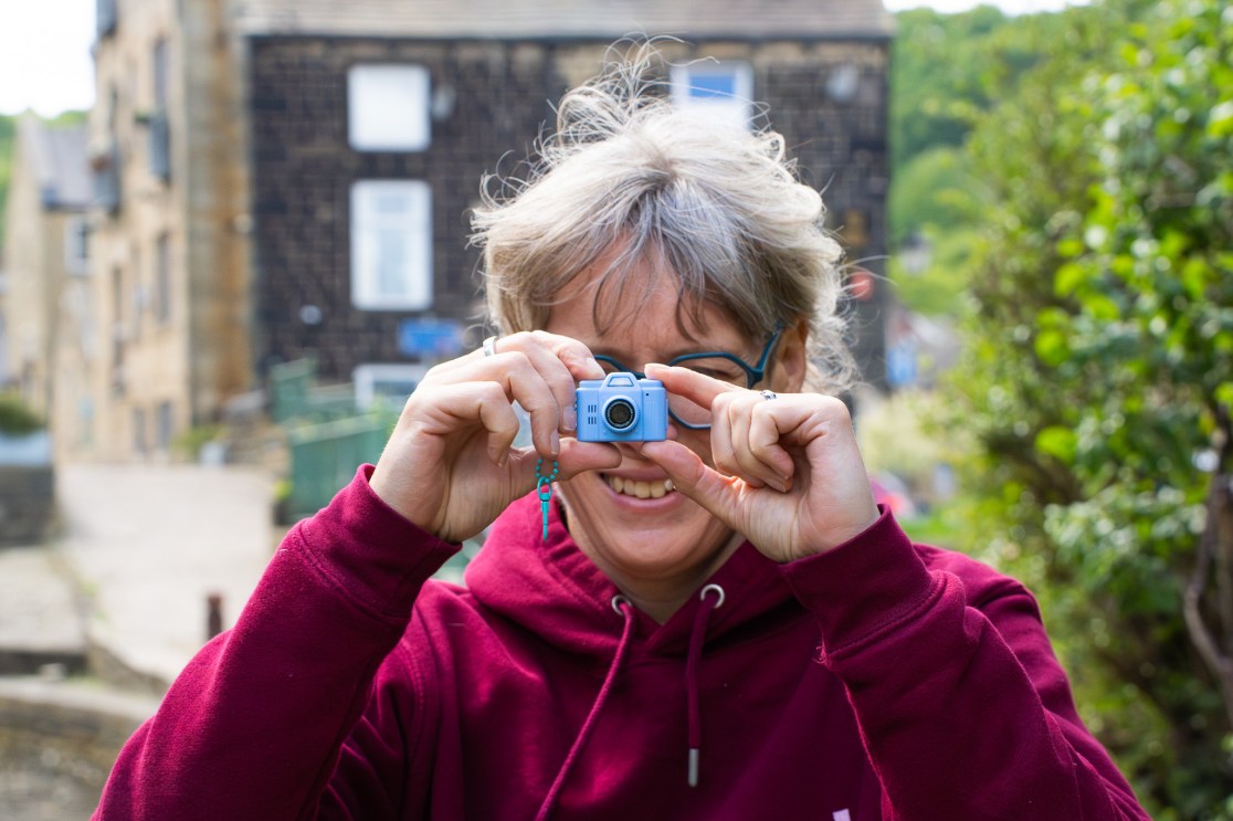 A smiling person holds a small blue digital camera up to their face, preparing to take a photo, with a background of buildings and greenery.