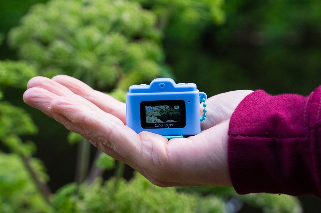 A blue mini digital camera held in a person's hand with greenery in the background.