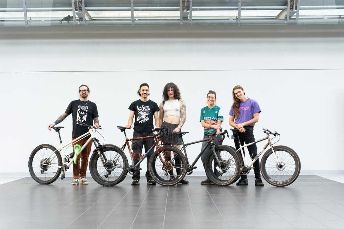 A group of five diverse individuals posing with their custom bikes in a modern indoor setting.