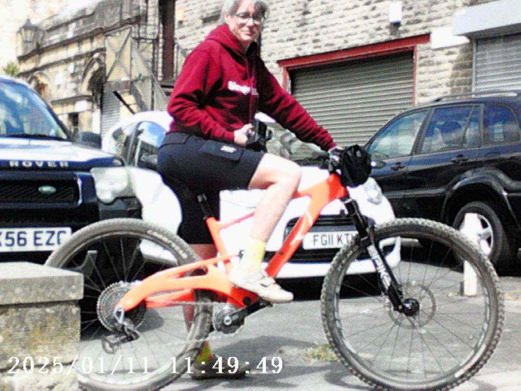 A person riding an orange ebike, wearing a red sweatshirt and shorts, with parked cars in the background.