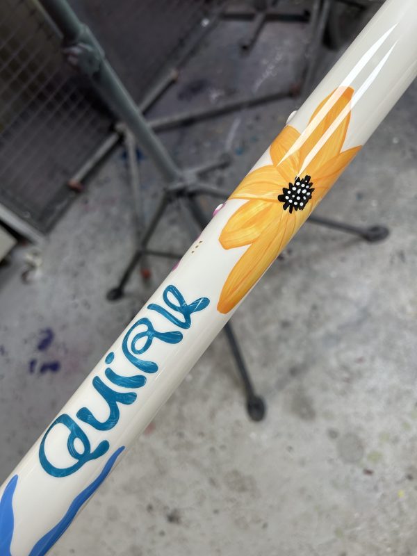 Close-up of a custom-painted bike frame featuring a vibrant yellow flower design and the word 'quirk' in blue lettering.