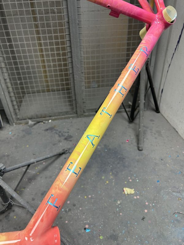 A close-up of a colorful bicycle frame showcasing a gradient paint finish from pink to yellow, with stylized lettering along the side in blue.