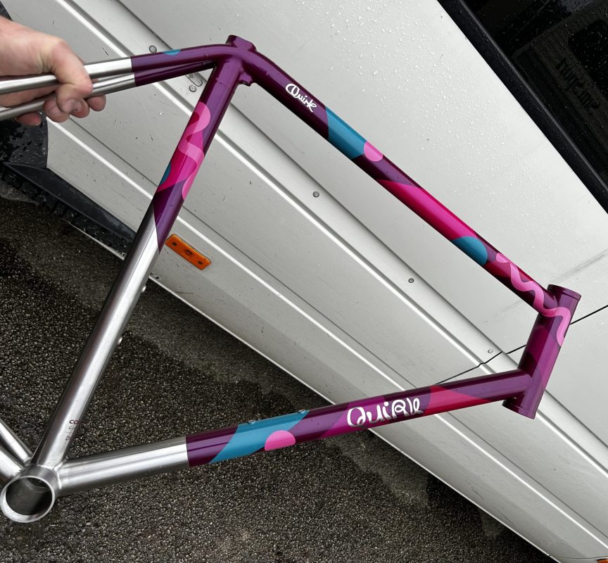 A close-up view of a vividly painted bicycle frame in purple and pink hues, with a blue accent, held by a person, against a backdrop of a vehicle.