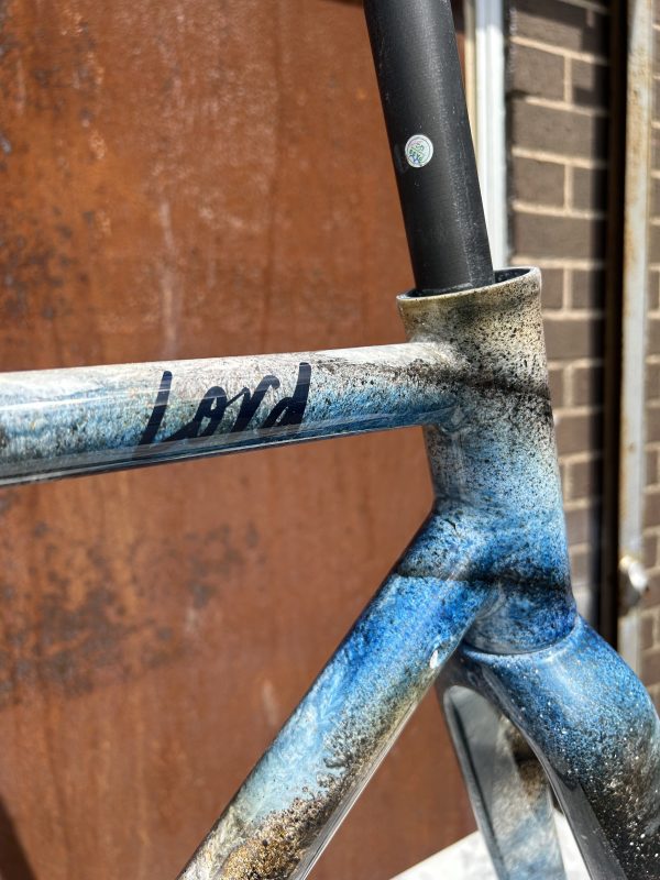 Close-up of a custom-painted bike frame featuring a blend of blue and black colors with the word 'Lord' visible on the top tube.