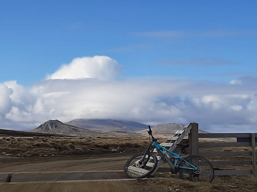 A mountain bike leaning against a wooden gate, with rolling hills and clouds in the background, depicting a scenic outdoor landscape.