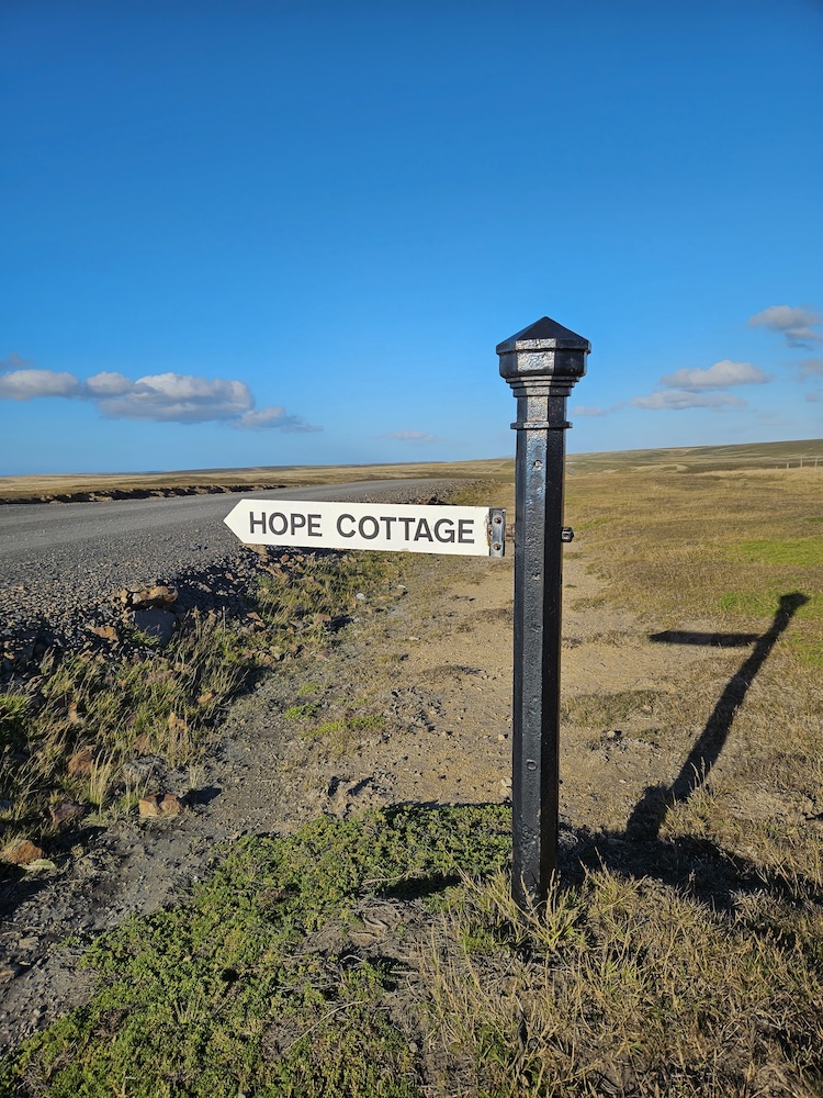 Signpost pointing toward Hope Cottage in a grassy landscape with a blue sky.