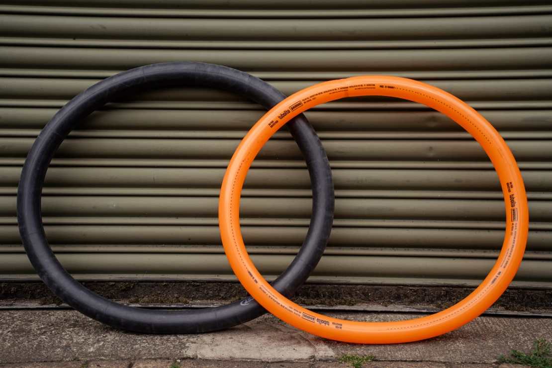 A comparison of a black butyl bicycle tube and a bright orange Tubolito tube, positioned side by side against a corrugated metal background.