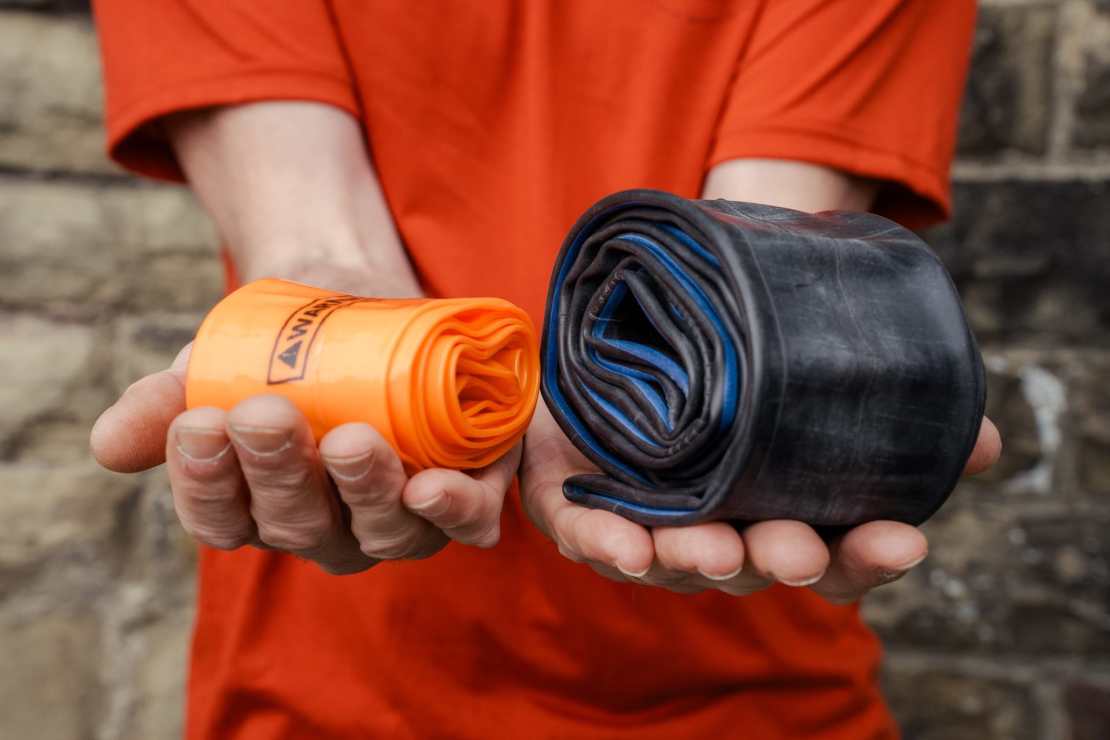 A person holding two bicycle inner tubes, one bright orange Tubolito tube on the left and a black butyl tube on the right, set against a stone wall.