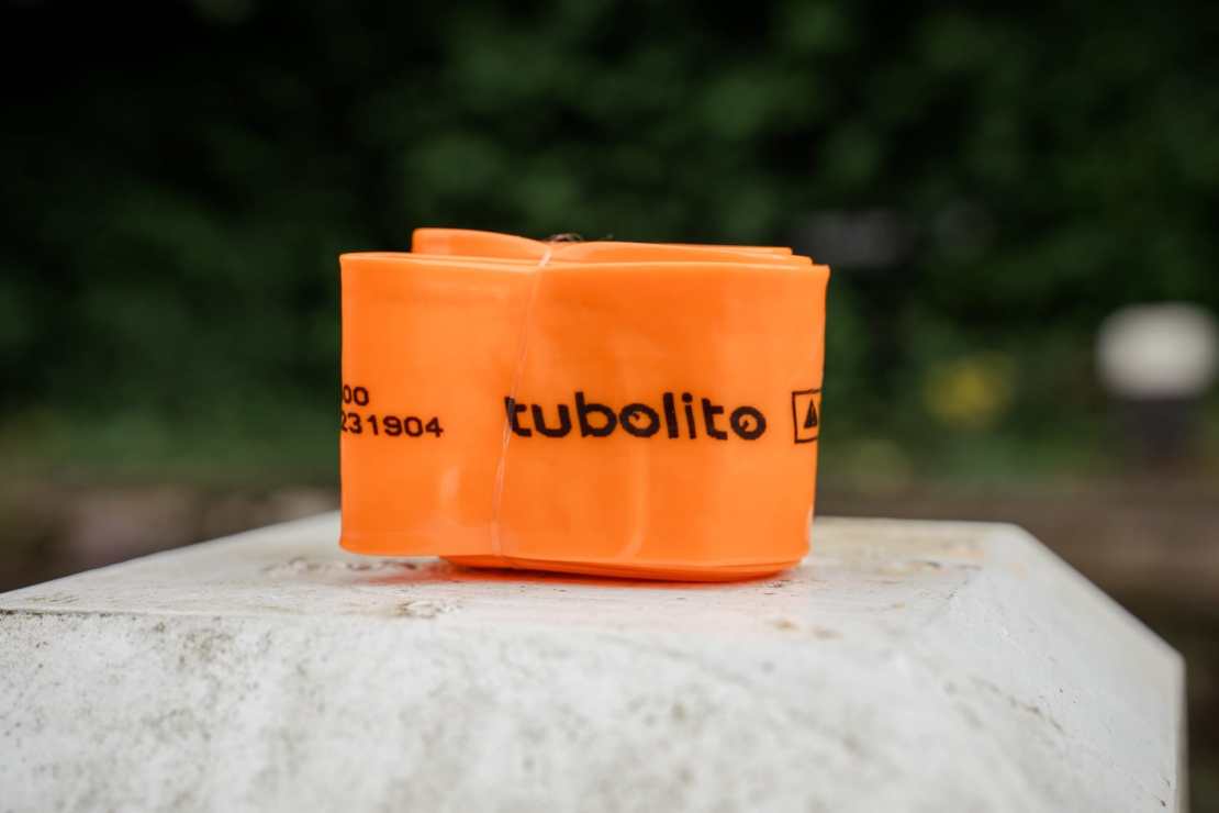 A compact, rolled Tubolito bicycle tube in bright orange color, featuring a black logo and text, displayed on a stone surface with a blurred natural background.