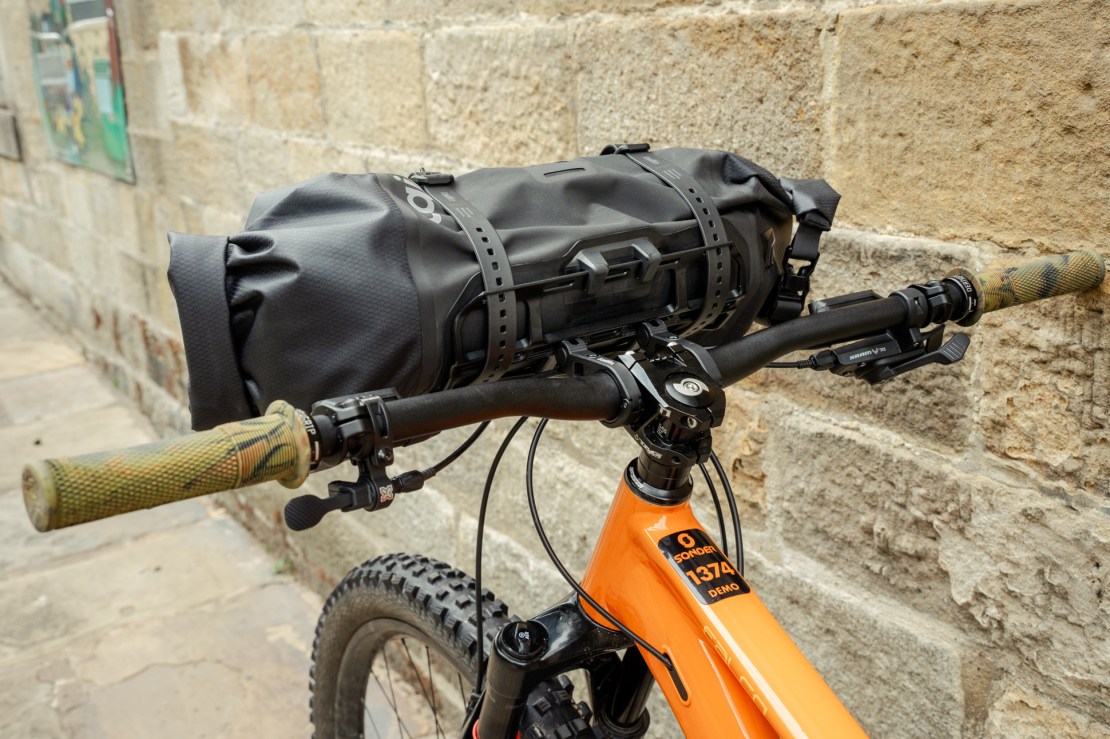 Close-up view of a bicycle handlebar featuring a black bag secured to the front, with patterned grips and a brick wall background.