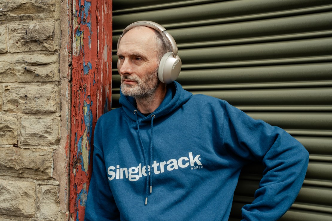 A man with a beard wearing a blue hoodie with 'Singletrack' branding, standing against a textured wall with a worn red frame and a green roller shutter background, while wearing silver headphones.