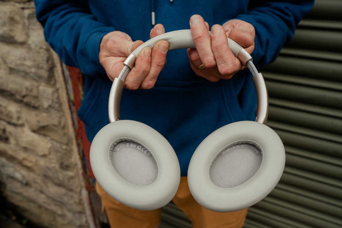 A person holding a pair of over-ear headphones with a soft, light-colored design, wearing a blue hoodie.