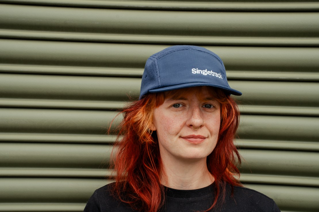 A person with red hair wearing a blue cap with the word 'Singletrack' embroidered on it, standing in front of a green corrugated background.