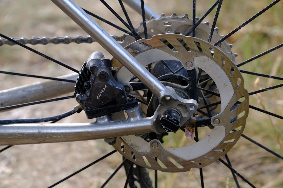 Close-up of a gravel bike's rear wheel showing the GRX brake caliper and disc brake, with a chain and spokes visible.