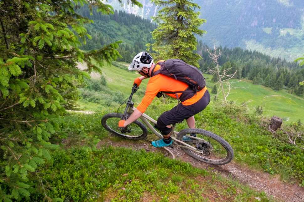 A mountain biker navigating a narrow trail surrounded by lush greenery and distant mountain views, wearing an orange jersey and protective gear.