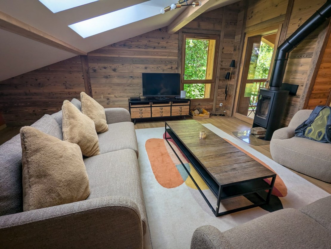 Cozy living room in a wooden chalet, featuring a sofa with plush pillows, a wooden coffee table, a television, and a wood stove, with large windows allowing natural light and a view of greenery.
