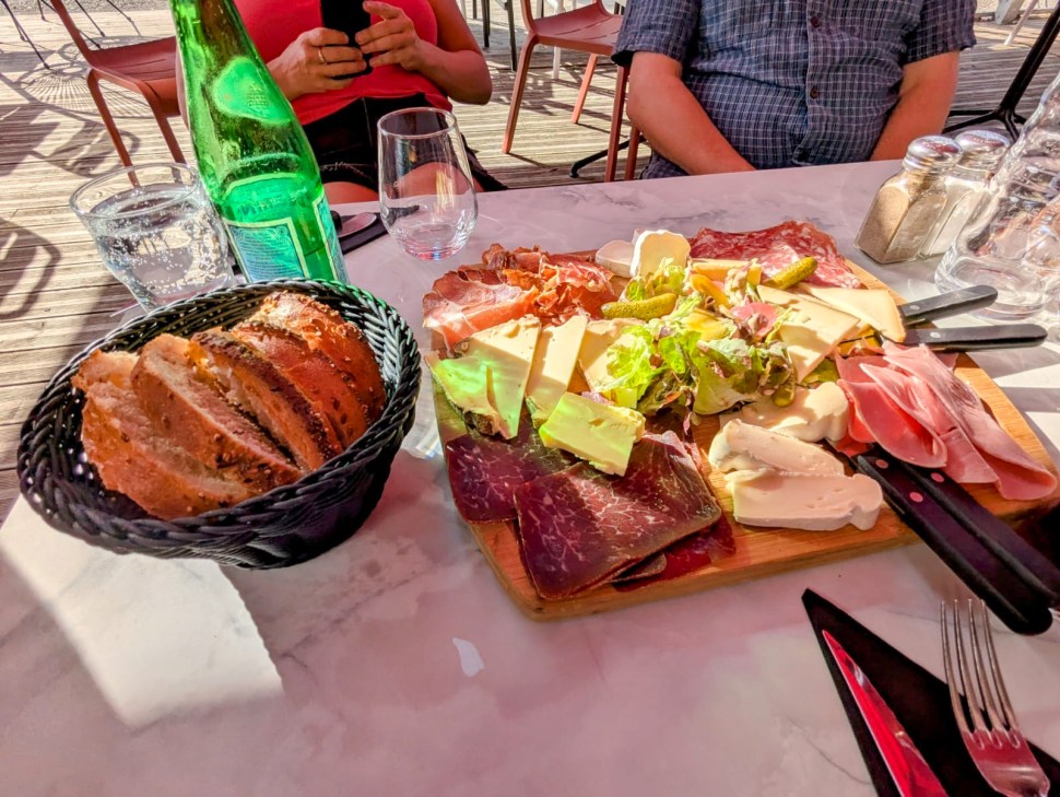 A wooden platter filled with a variety of cheeses, cured meats, pickles, and fresh greens, with a basket of sliced bread and a sparkling water bottle in the foreground.