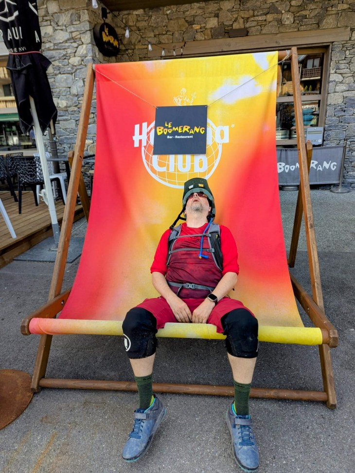 A cyclist resting on a large deck chair outside a bar-restaurant called Le Boomerang in Morzine, France, wearing a helmet and sports gear.