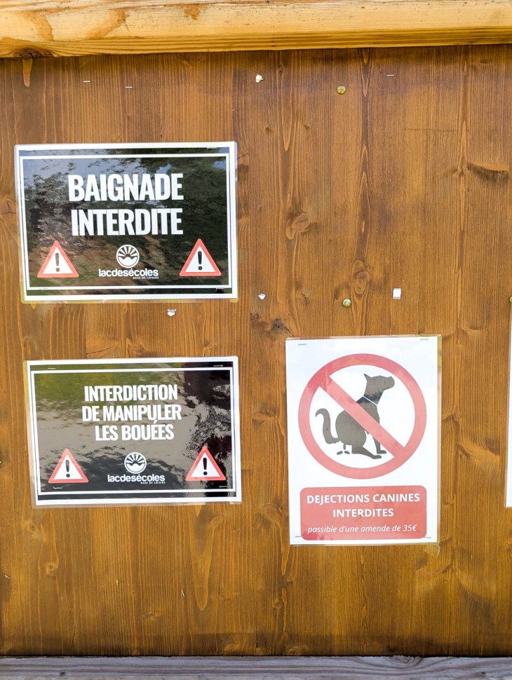 A wooden sign displaying three warnings in French: 'Baignade interdite' (swimming prohibited), 'Interdiction de manipuler les bouées' (do not handle the buoys), and 'Déjections canines interdites' (no dog waste allowed) with a fine notice.