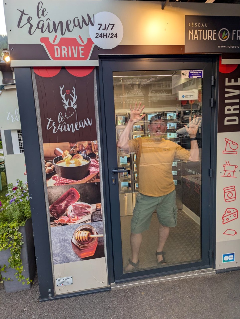 A person playfully pretending to be trapped inside a food vending machine with a sign indicating it is a 24/7 drive-thru service in Les Gets, France.
