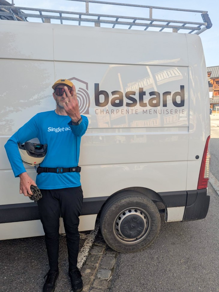 A person wearing a blue long-sleeve shirt and helmet stands next to a white van with the logo 'bastard' on the side, gesturing peace signs.