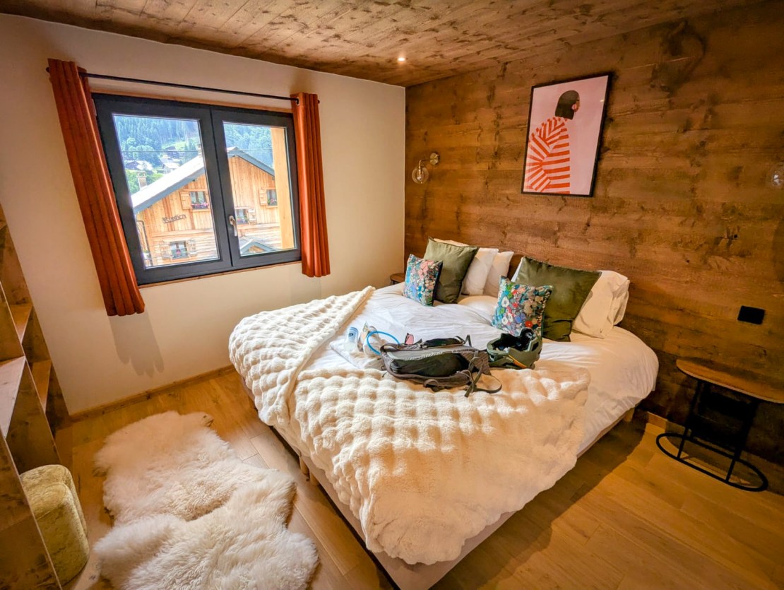Cozy bedroom with a large bed, decorative pillows, and a warm color scheme, featuring wooden walls and a window overlooking a mountain village.  Bike Morzine