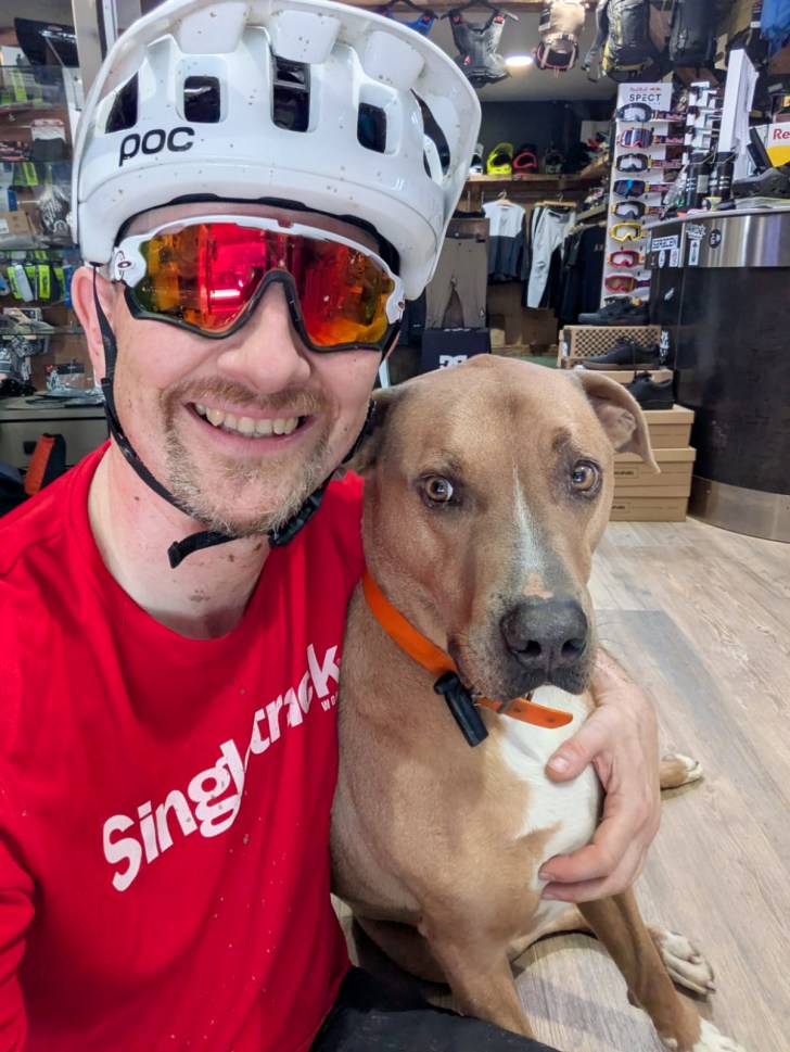 A smiling person wearing a white helmet and sunglasses poses with a brown dog in a bike shop setting.