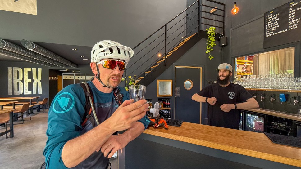 A mountain biker in a helmet holding a glass while talking at the IBEX brewery, with a bar and a staff member visible in the background.