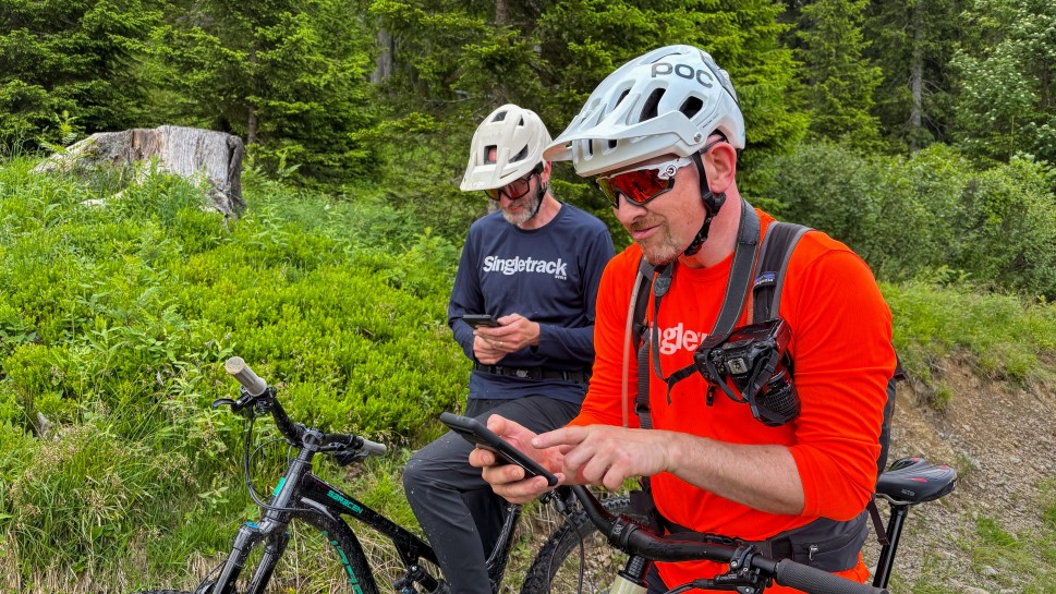 Two mountain bikers in a forested area, one checking their phone while the other looks on. They are wearing helmets and cycling gear.