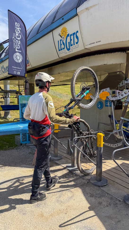 A mountain biker preparing to load their bike onto a lift at Les Gets in the French Alps, with signage visible in the background.