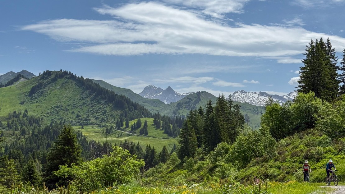 Mountain landscape in Les Gets, France, featuring lush green hills, snow-capped peaks, and two cyclists riding on a trail.