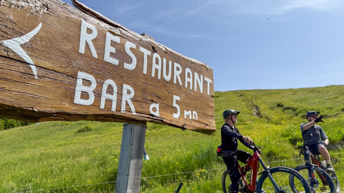 A wooden sign indicating a restaurant and bar, with two cyclists in the foreground, surrounded by green fields under a clear blue sky.