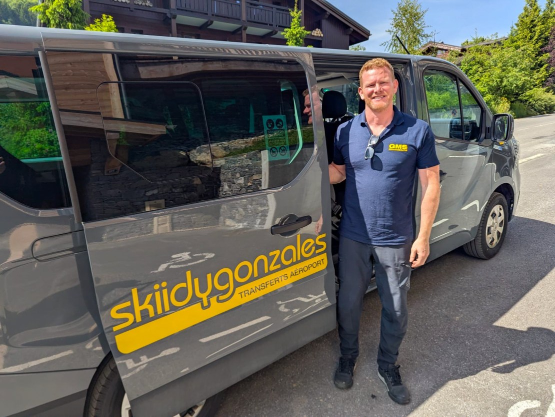 A driver stands next to a Skiidy Gonzales transfer van, ready to assist with airport transportation for mountain bikers.