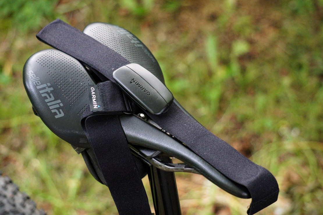 A Garmin heart rate monitor attached to a Selle Italia saddle on a mountain bike, set against a blurred greenery background.
