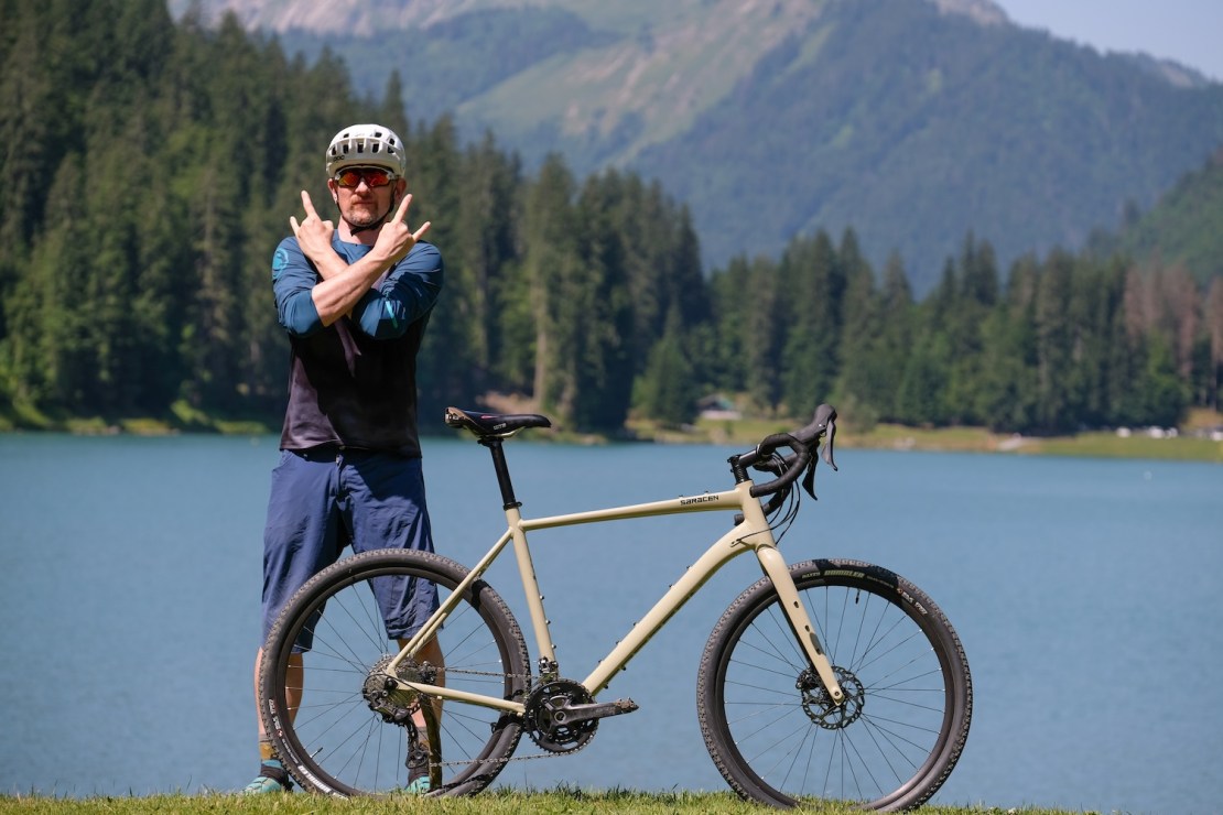 A person with a helmet and sunglasses poses with a gravel bike by a lake surrounded by mountains and trees.