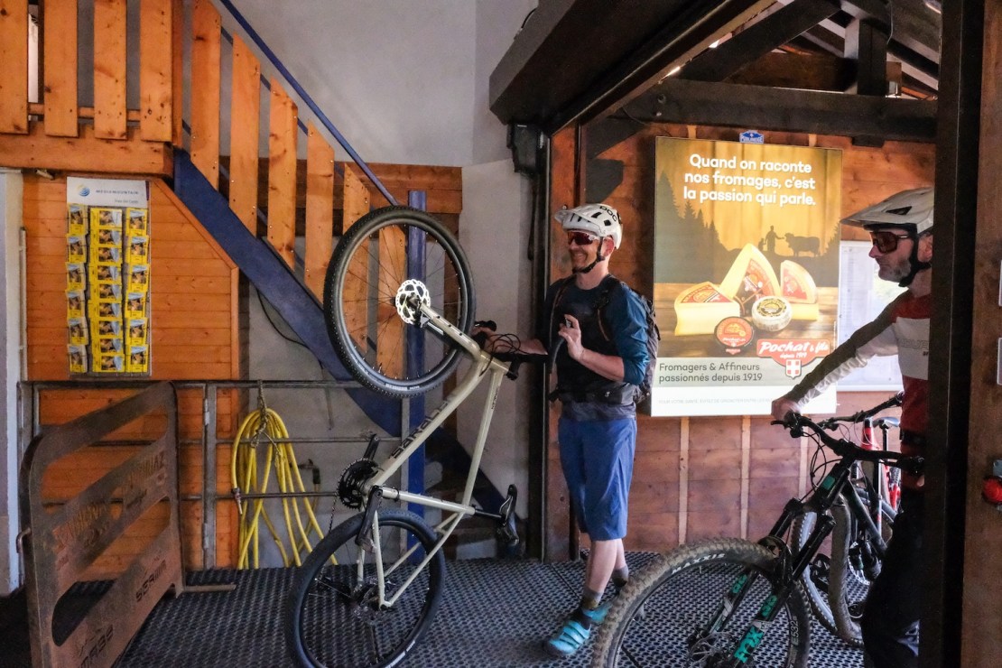 A cyclist holding a gravel bike with one wheel elevated in a bike lift area, while another cyclist prepares to enter. Wooden stairs and a promotional poster on the wall are visible in the background.