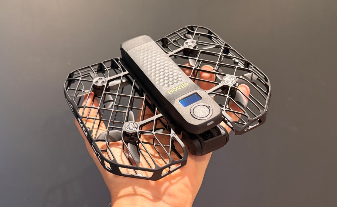 A close-up image of a hand holding a compact drone with a protective grid frame, featuring a digital display and a textured surface.
