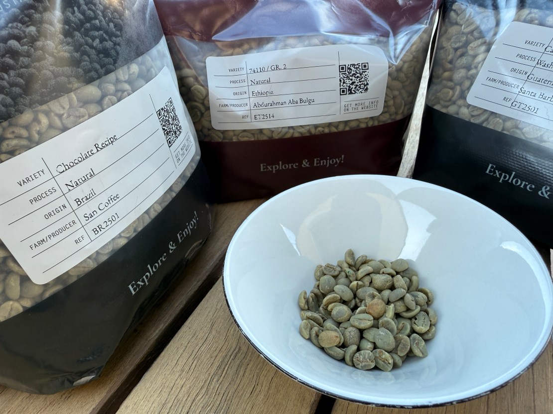 A bowl filled with green coffee beans, surrounded by bags of coffee labeled with different varieties and origins, set on a wooden surface.