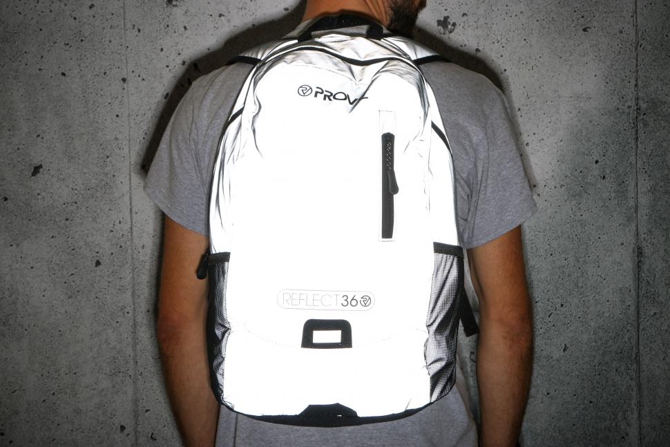 A person wearing a gray t-shirt is standing against a concrete wall, showcasing a reflective white backpack with the brand 'PROV' and the label 'REFLECT 36'.