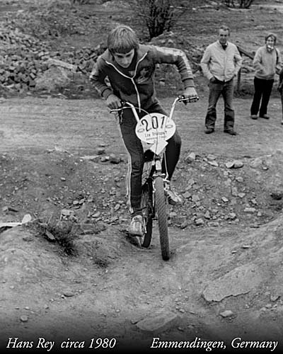 A black and white photo of a young boy, Hans Rey, riding a bicycle over rough terrain in Emmendingen, Germany, circa 1980. He is focused on navigating the obstacle while wearing a tracksuit.