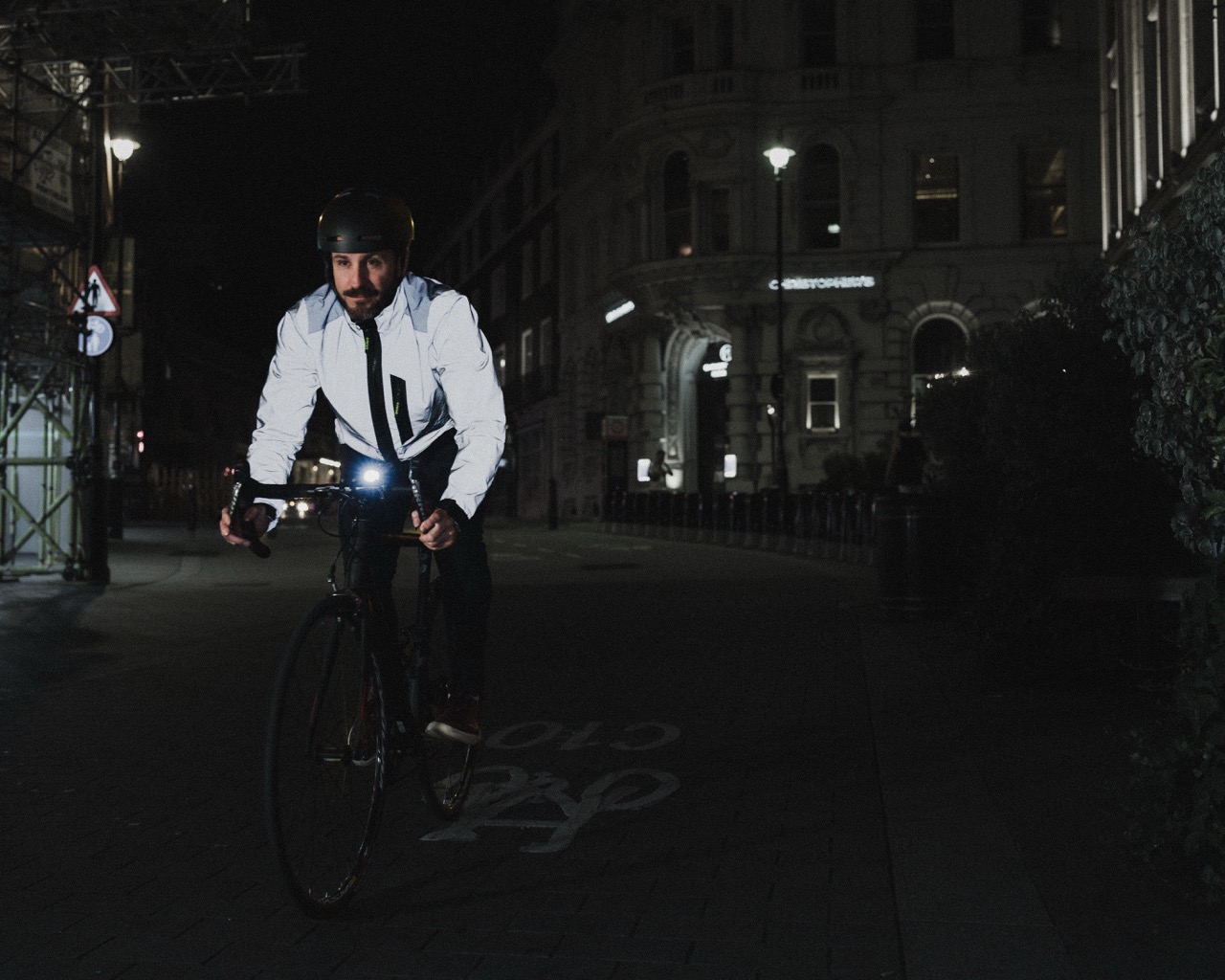 A cyclist wearing a reflective jacket and helmet rides a bicycle at night in an urban setting.