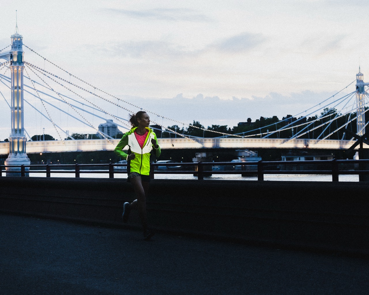A woman jogging along a riverbank at dusk, wearing a bright reflective cycling jacket, with a bridge and city skyline in the background.