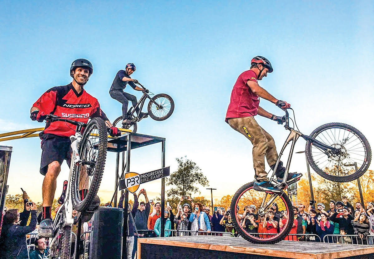 Three riders perform tricks on bicycles at a bike trials event, with a crowd watching and a clear blue sky in the background.