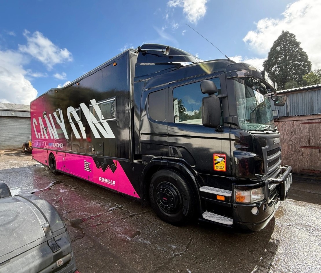 A large Scania P310 Motorhome Race Truck with a black and pink design, parked with cloudy skies in the background.