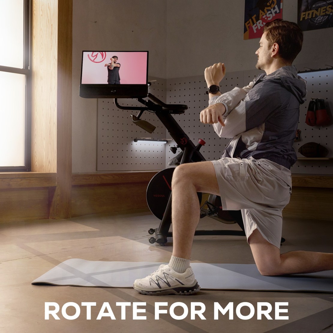 A person performing a workout on a mat, facing a rotating display screen of the Yesoul G1 M Plus indoor bike in a well-lit room.