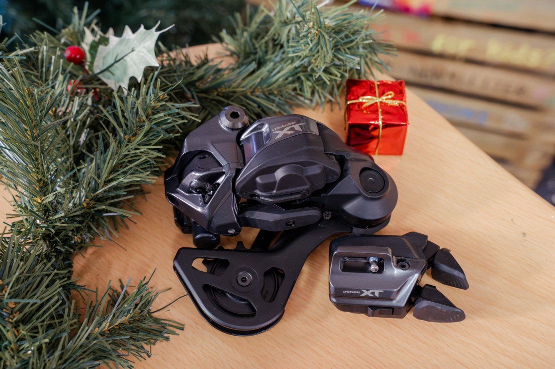 Close-up of Shimano XT Di2 rear derailleur and shifter on a wooden surface, surrounded by pine branches and a small red gift box.