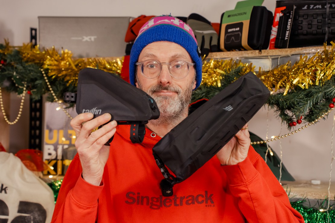 A person wearing a colorful beanie and red hoodie holds two different bike bags against a festive backdrop with decorations.